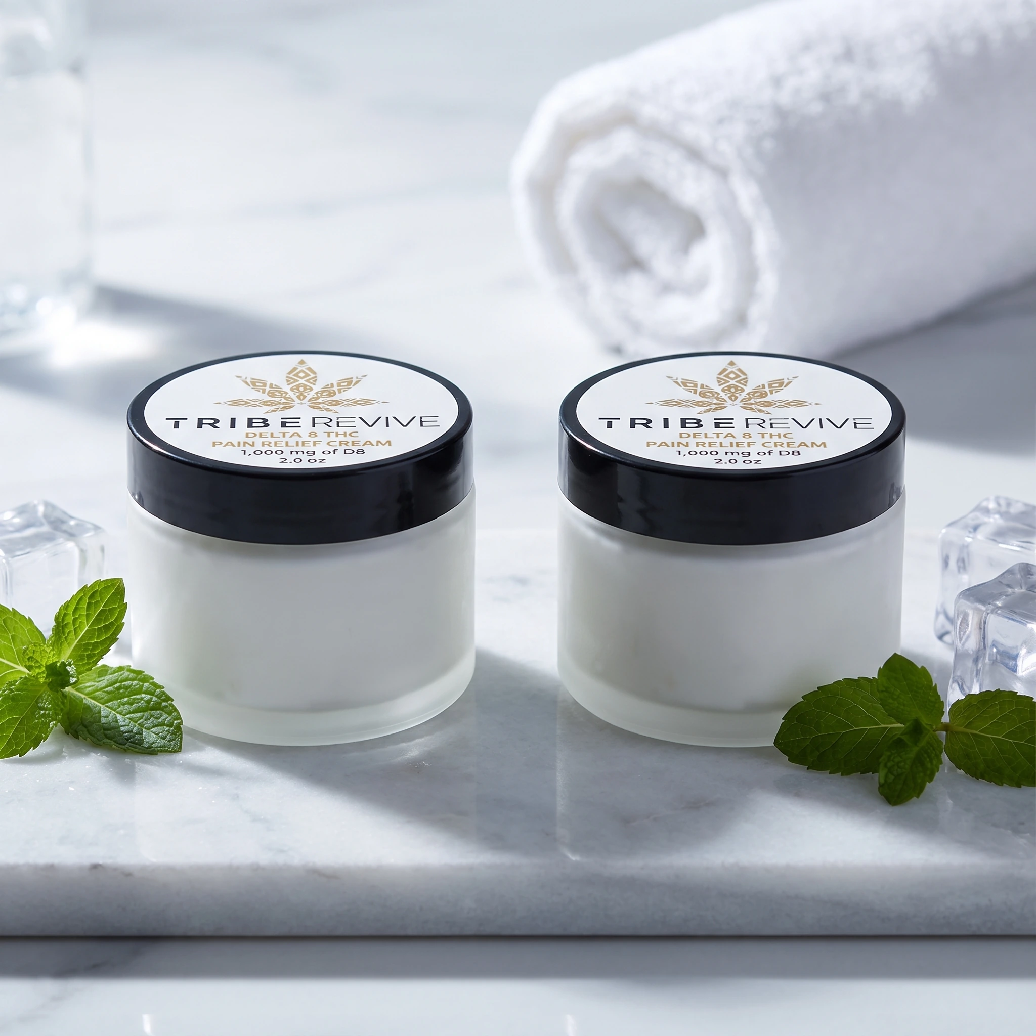 Two jars of TribeTokes TribeRevive Delta 8 THC Pain Relief Cream on white marble with fresh mint and ice cubes, and a rolled towel behind