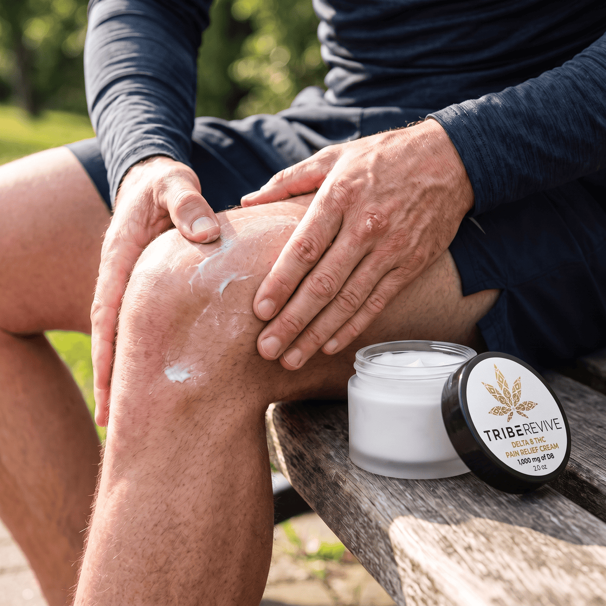 Man in athletic wear rubbing TribeRevive Delta 8 THC Pain Relief Cream into his knee outdoors, the open jar on a wooden bench beside him