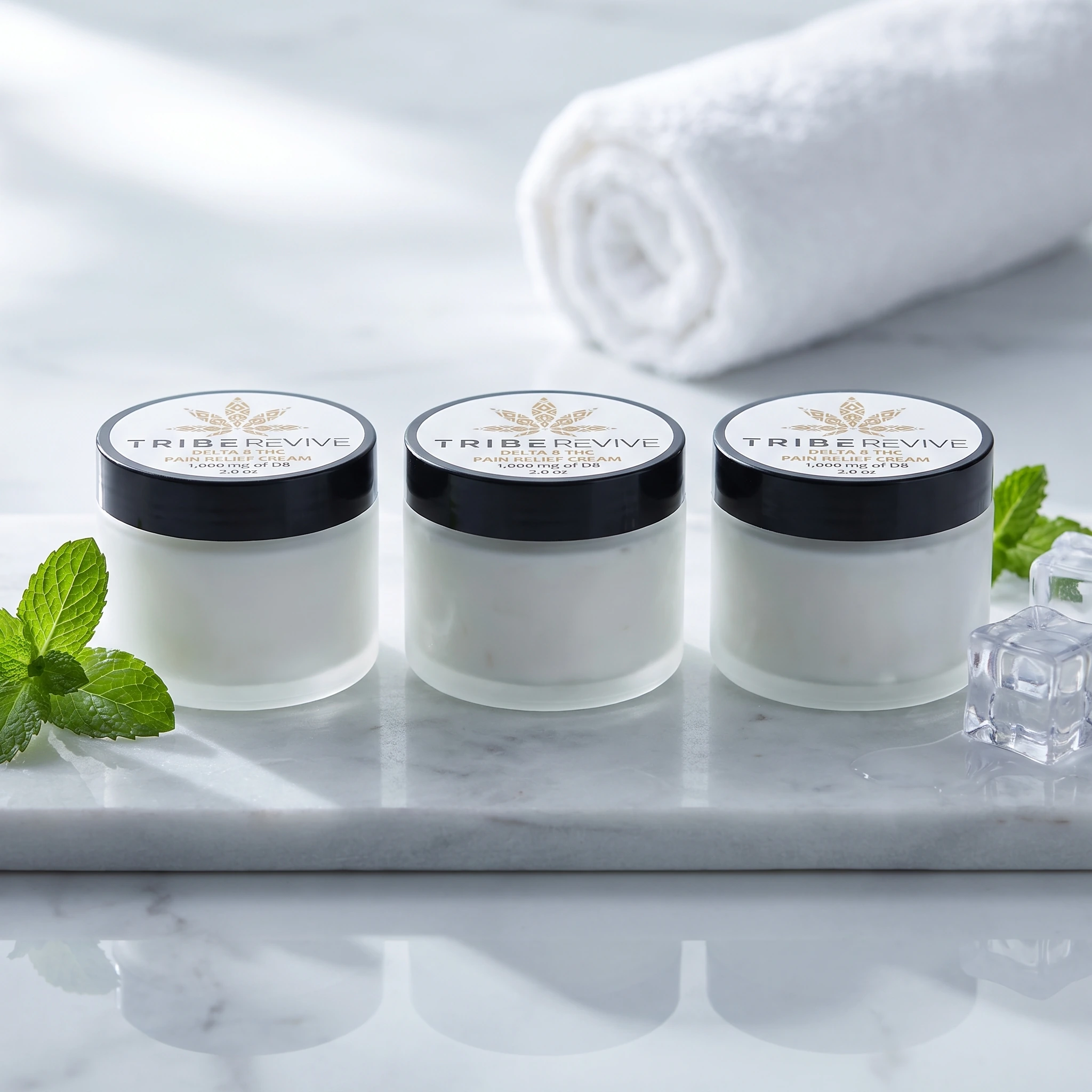 Three jars of TribeTokes TribeRevive Delta 8 THC Pain Relief Cream on white marble with fresh mint and ice cubes, and a rolled towel behind