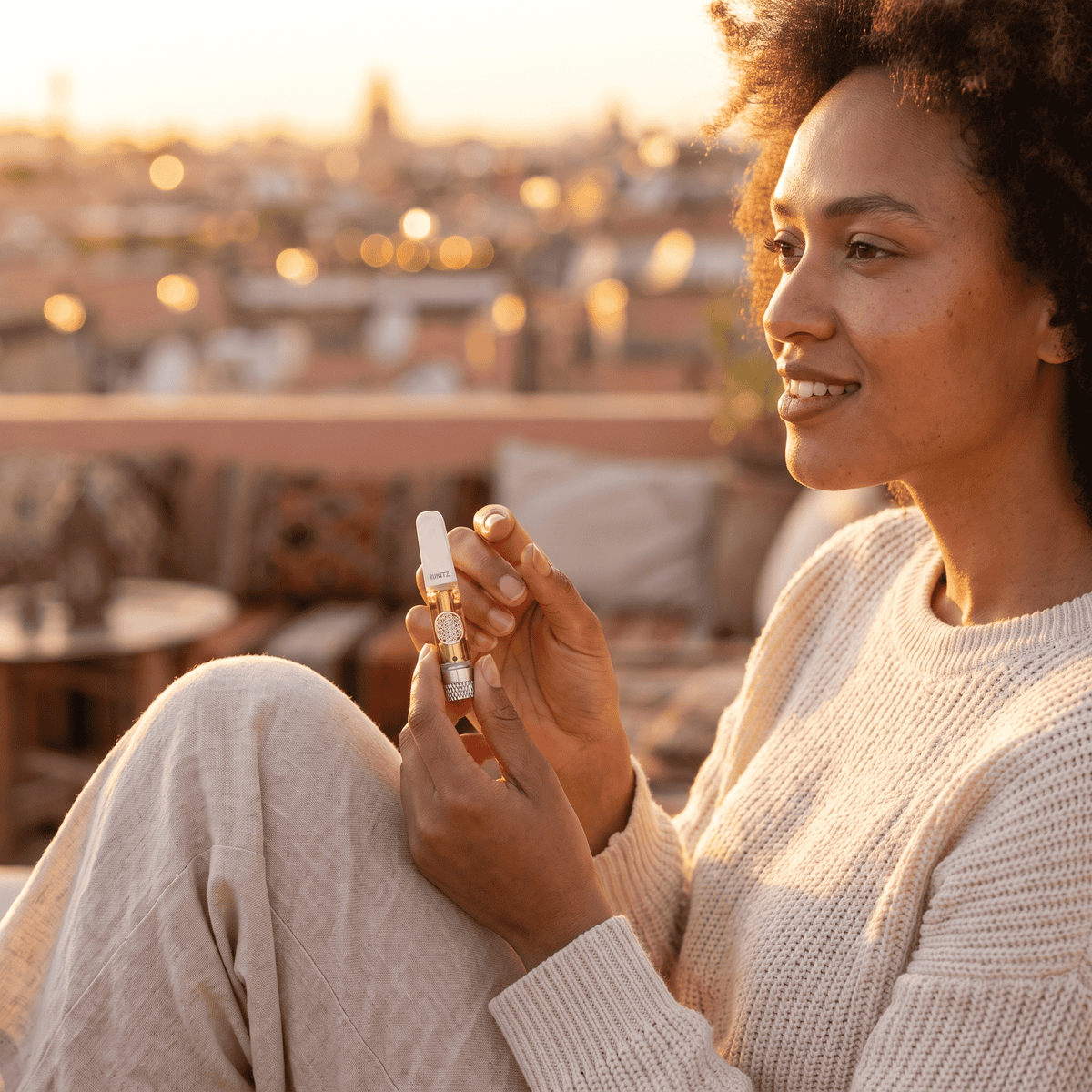Woman in a cream knit set relaxing on a rooftop at golden hour, holding a TribeTokes THCa Live Resin cartridge