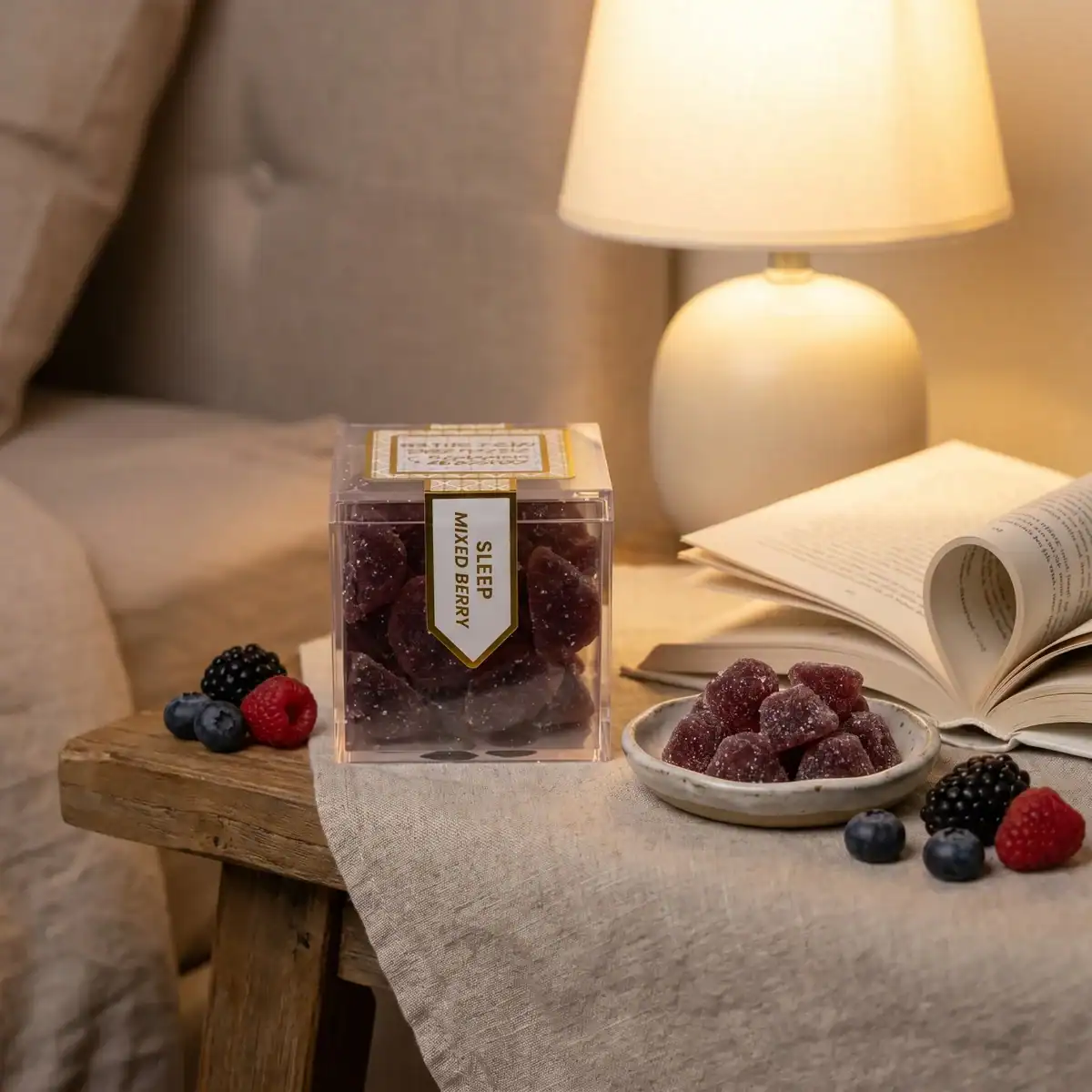 TribeTokes Sleep Mixed Berry gummies on a nightstand beside an open book and a lit ceramic lamp in warm evening light