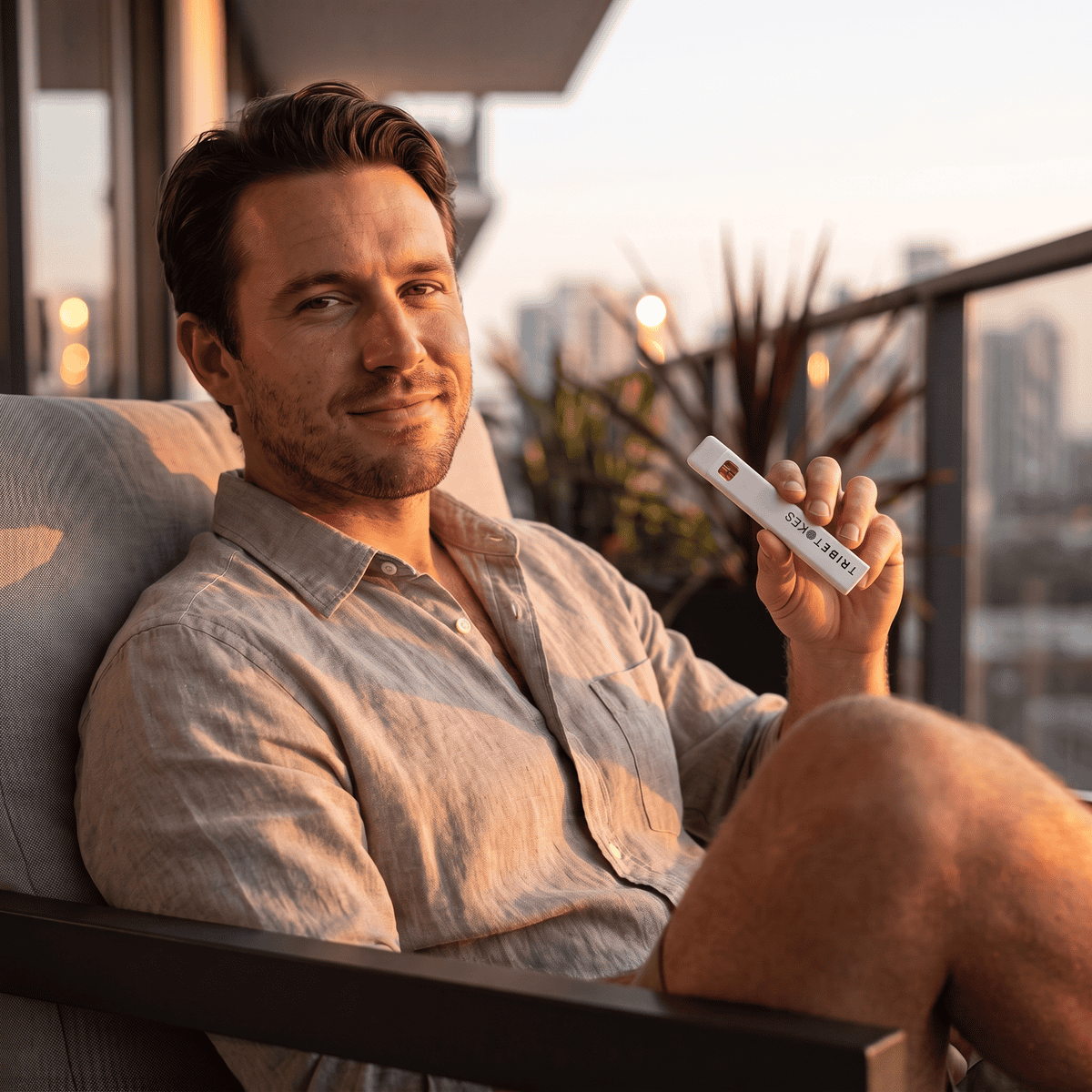 Man in a linen shirt relaxing on a balcony sofa at golden hour, holding a TribeTokes THCa Live Resin disposable vape with the city skyline behind him