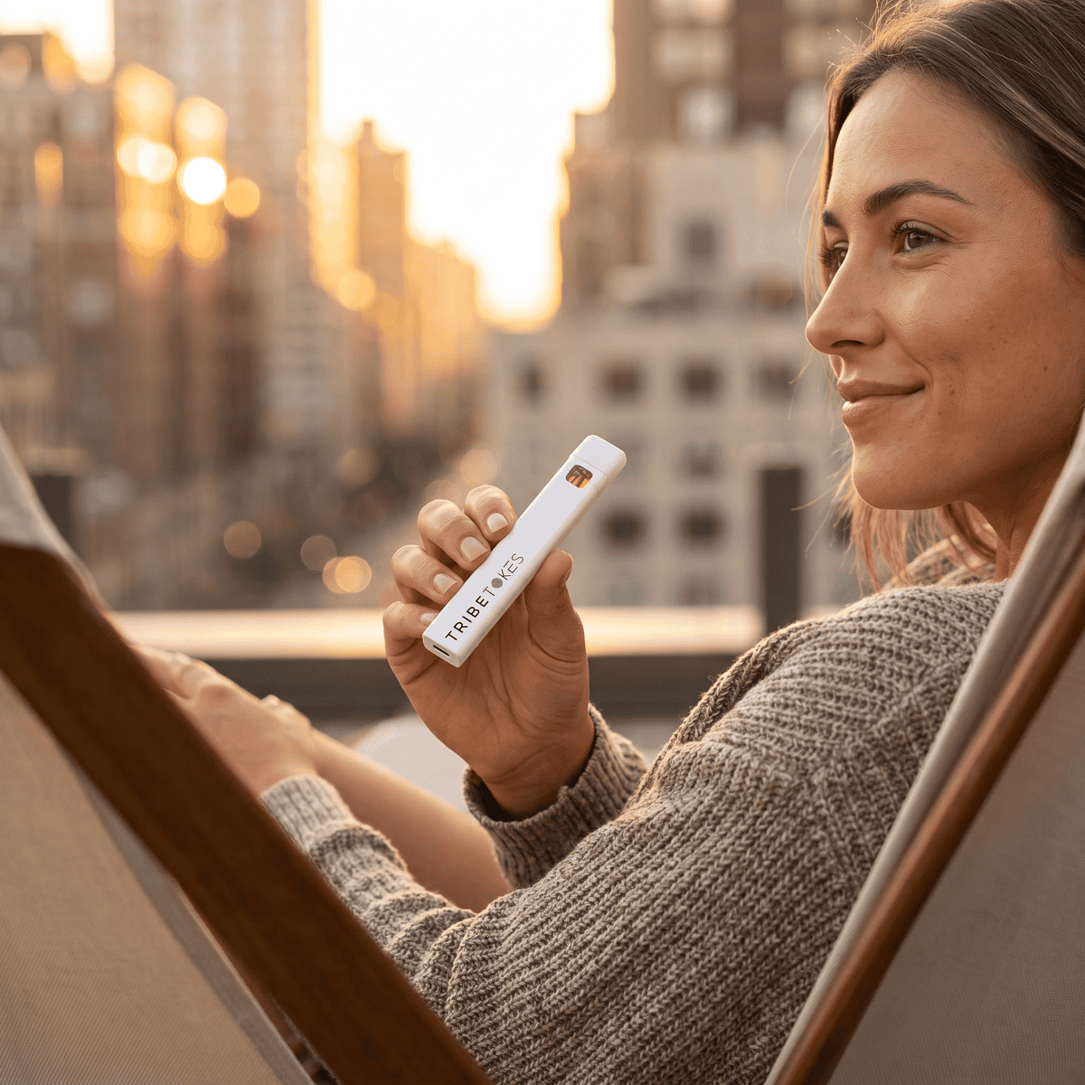 Woman in a knit sweater reclining on a rooftop chair at sunset, holding a TribeTokes THCa Live Resin disposable vape with the city behind her
