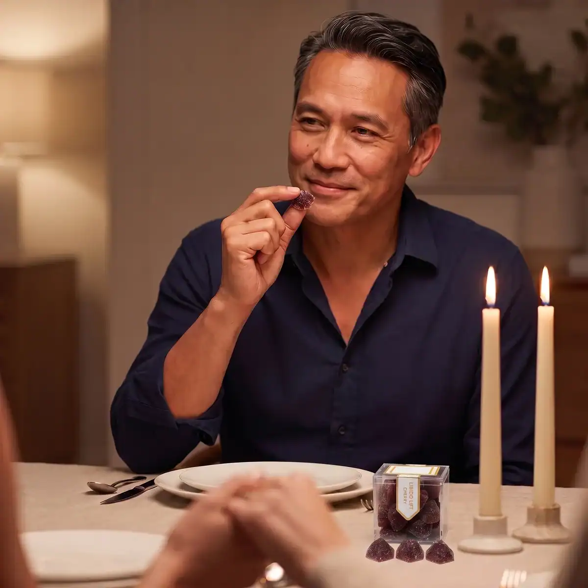Man at a candlelit dinner smiling across the table, holding a TribeTokes CBD Libido Lift Cherry gummy with the cube box beside the candles