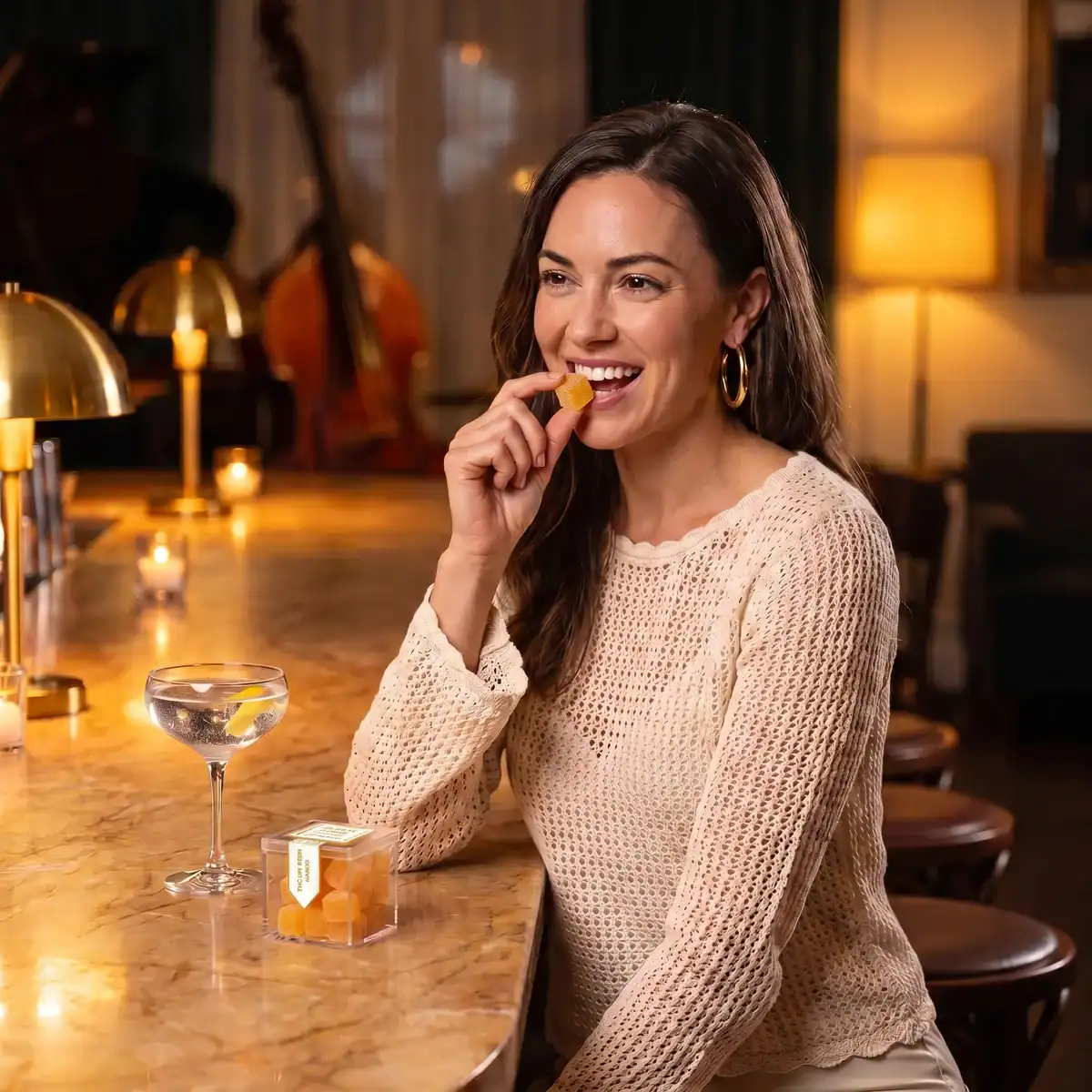 Woman in a cream knit top smiling at a candlelit jazz bar, eating a TribeTokes THC Live Resin Mango gummy with the cube box on the marble counter