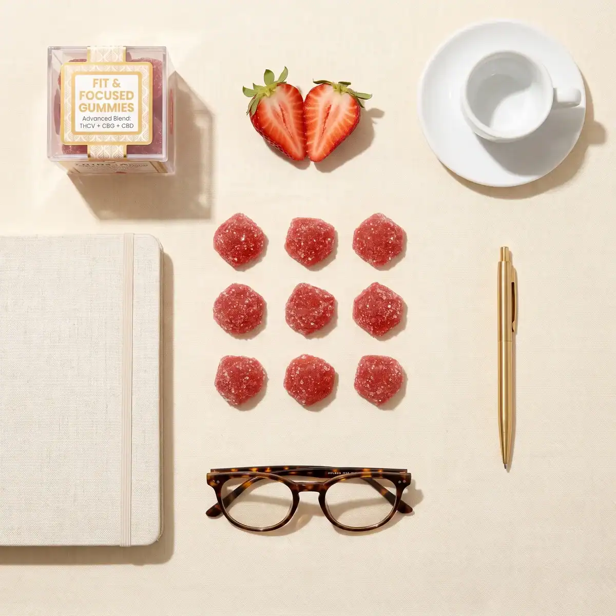 Flat lay of TribeTokes Fit & Focused Strawberry gummies arranged with an open notebook, tortoiseshell glasses, a gold pen, an espresso cup, and a sliced strawberry on cream linen