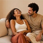 Man and woman laughing together on a cream couch in natural light, representing cannabis and sexual wellness