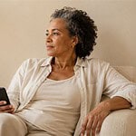 Middle-aged person relaxing in a comfortable armchair holding a phone in soft natural daylight with warm neutral home décor and a calm thoughtful expression.