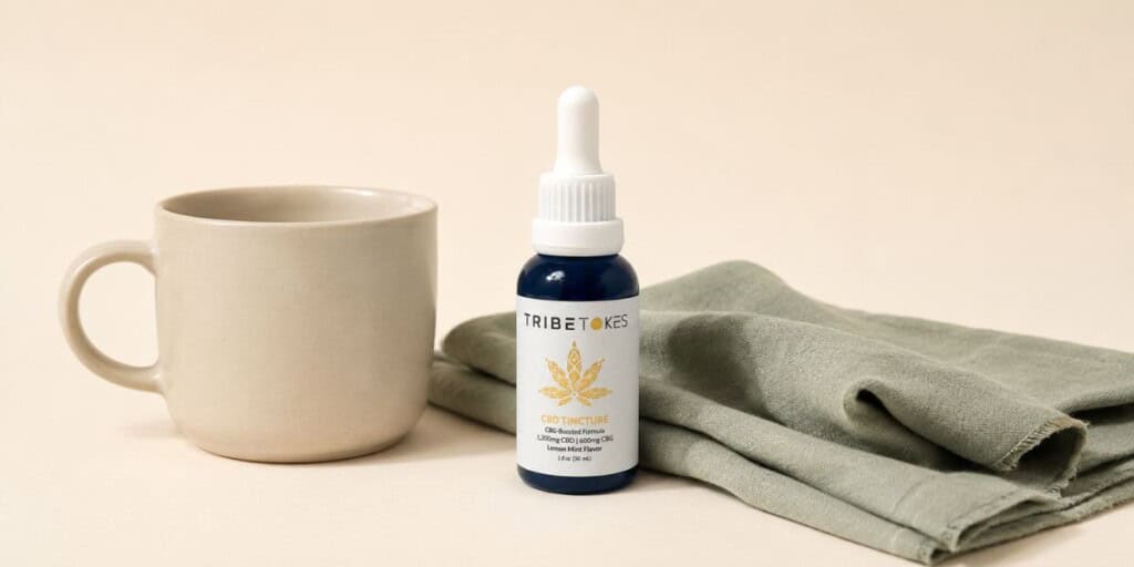 TribeTokes CBD tincture bottle beside a matte ceramic mug and a folded sage linen napkin on a cream background with soft natural light creating a calm everyday wellness scene.