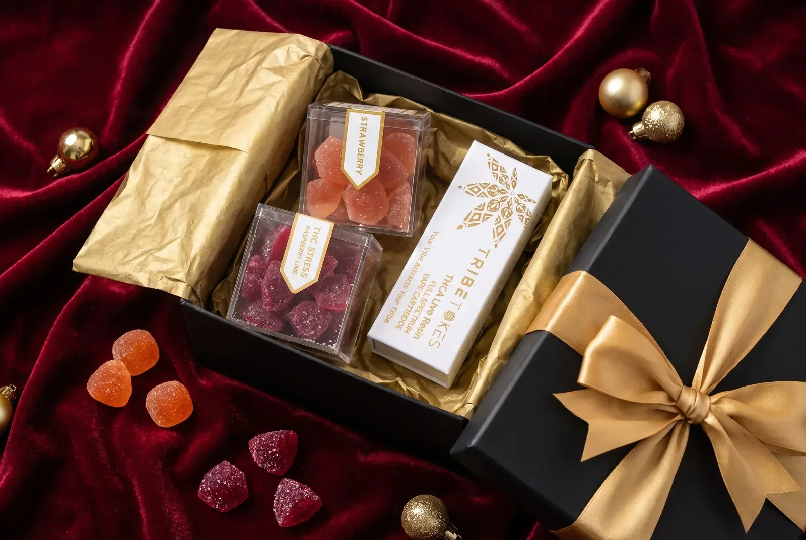Open black gift box with gold ribbon holding two boxes of TribeTokes gummies and a THCa live resin vape cartridge