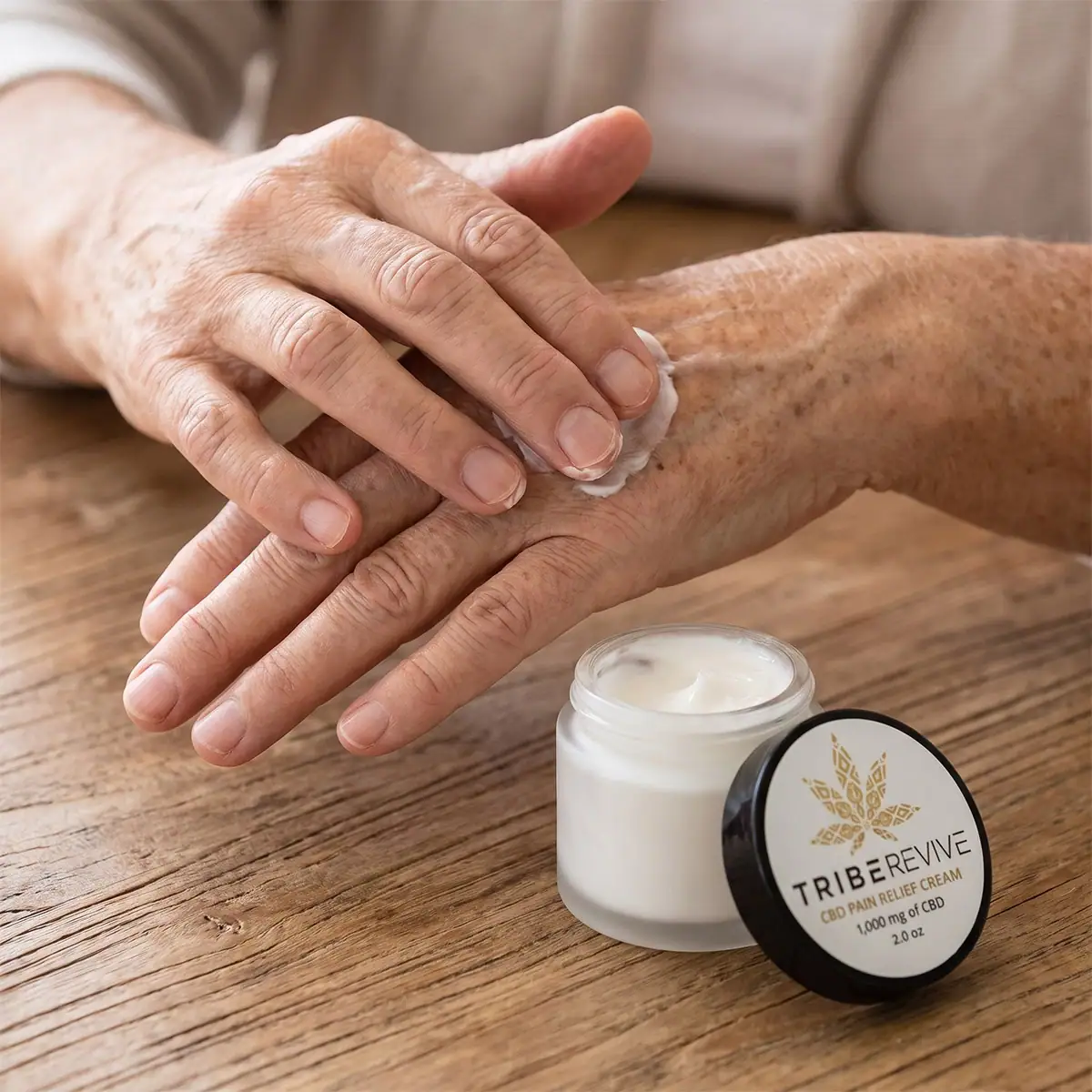 Older man massaging TribeRevive CBD Pain Relief Cream into the back of his hand at a wooden table, the open jar beside him