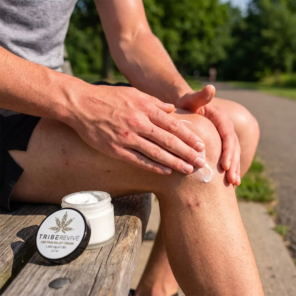 Man in athletic wear rubbing TribeRevive CBD Pain Relief Cream into his knee outdoors, the open jar on a wooden bench beside him