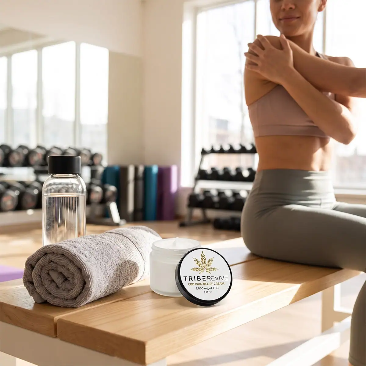 Woman stretching in a sunlit gym with a rolled towel and water bottle on the bench beside an open jar of TribeRevive CBD Pain Relief Cream