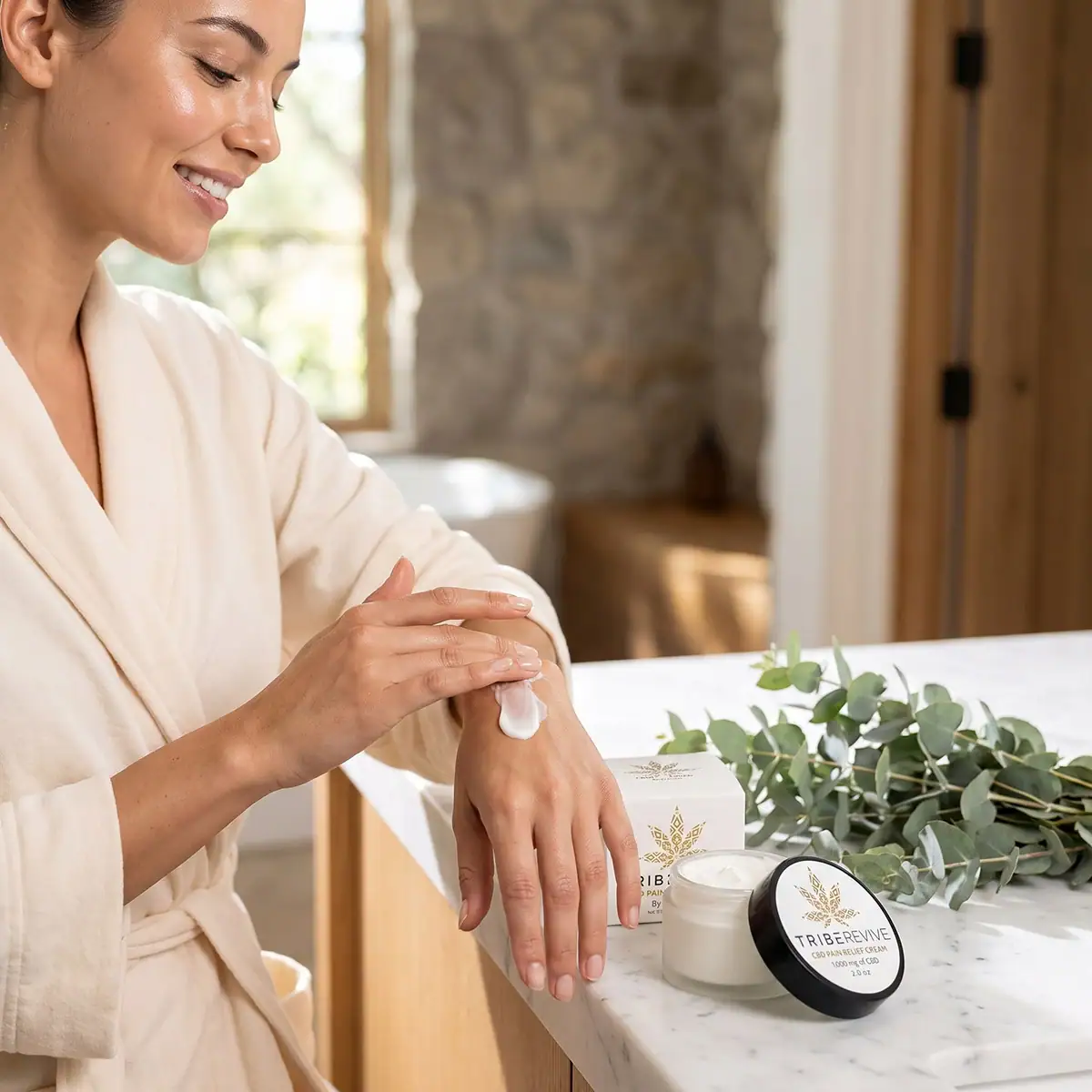 Woman in a robe smoothing TribeRevive CBD Pain Relief Cream onto the back of her hand at a marble spa vanity with eucalyptus and the jar beside her