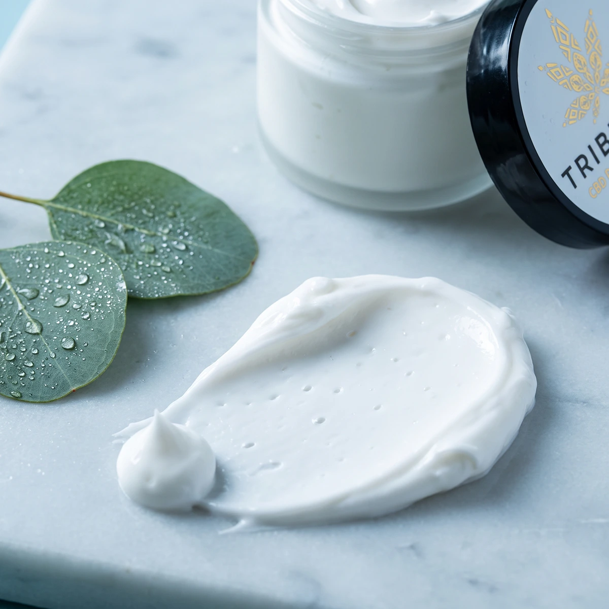 A swipe of TribeRevive CBD Pain Relief Cream on marble beside dewy eucalyptus leaves, with the open jar behind it