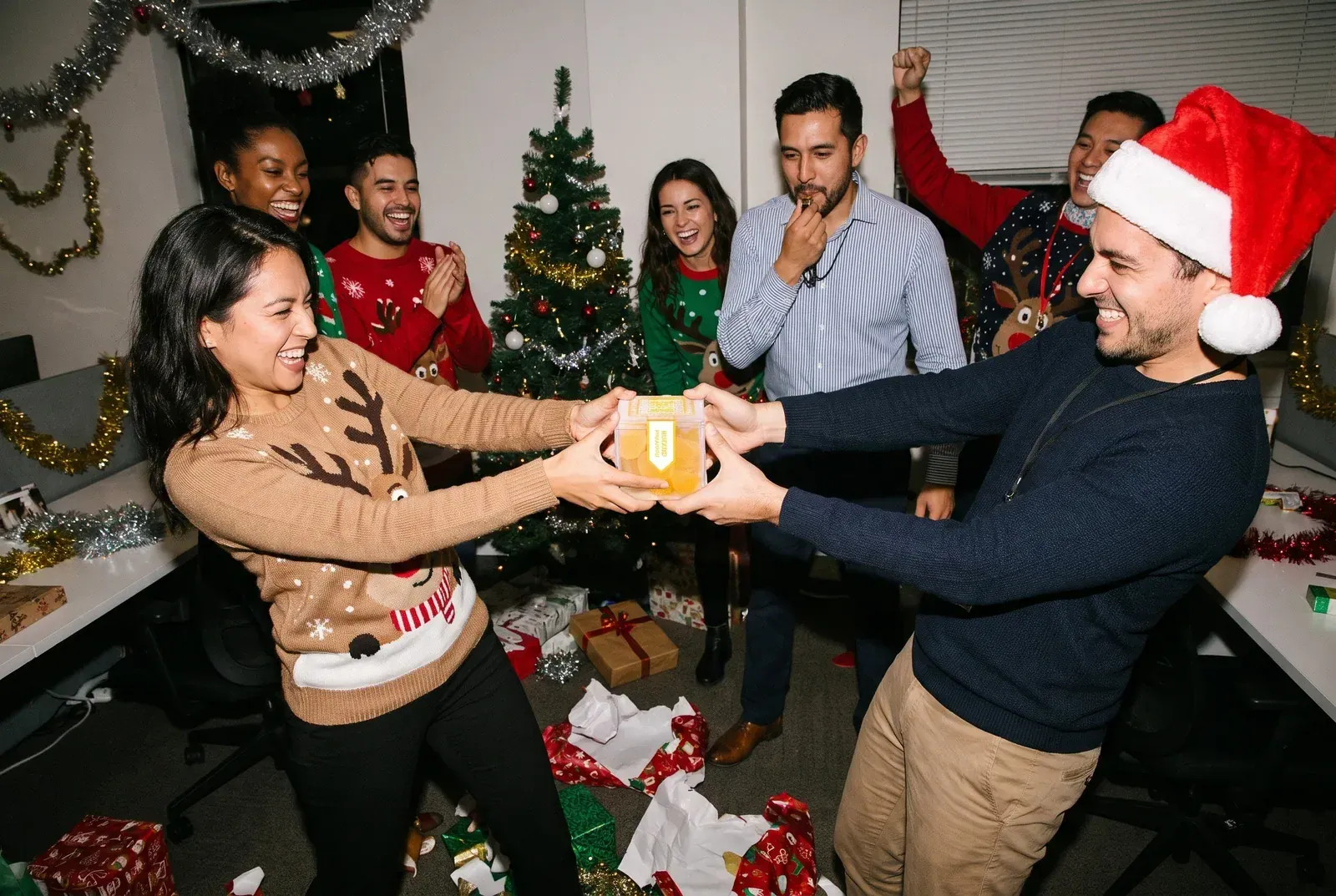 Two coworkers in holiday sweaters playfully tugging on a box of TribeTokes gummies during an office gift exchange